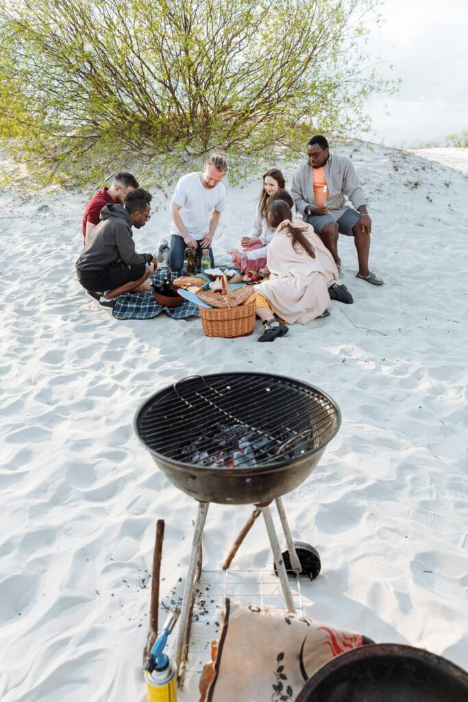 A group of adults have a picnic on a sandy beach near a grill, enjoying a sunny day.
